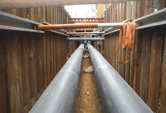 Twin district heating pipes running along a trench lined with trench sheets and hydraulic waler frames, with a lifting strap on a strut
