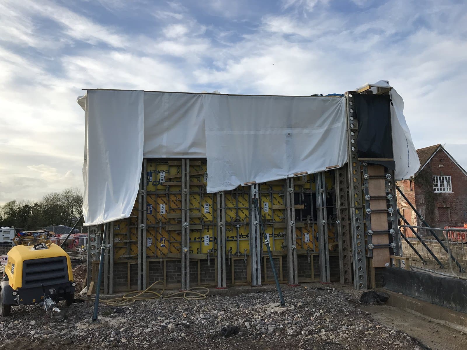 Frami formwork with Slimshore soldiers partly covered in white weather sheeting beside a compressor on the Wiltshire site