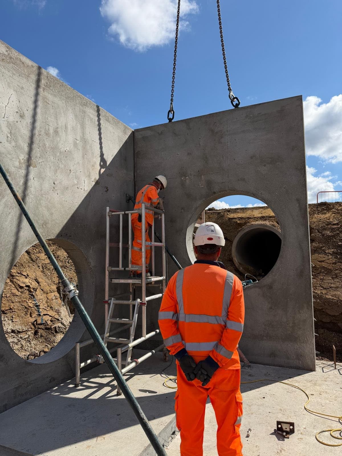 Precast manhole wall panel on crane chains being placed beside a standing panel braced by a push-pull prop