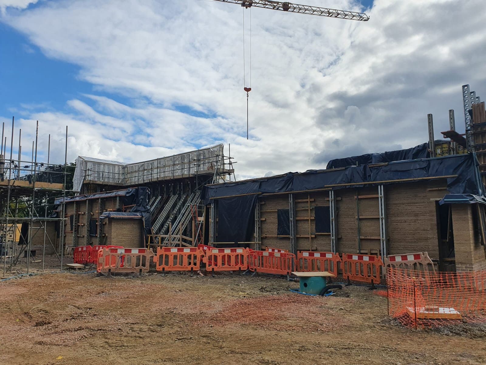 Rammed earth walls rising on the Wiltshire site behind pedestrian barriers, with Slimshore soldiers, black protective sheeting and a tower crane