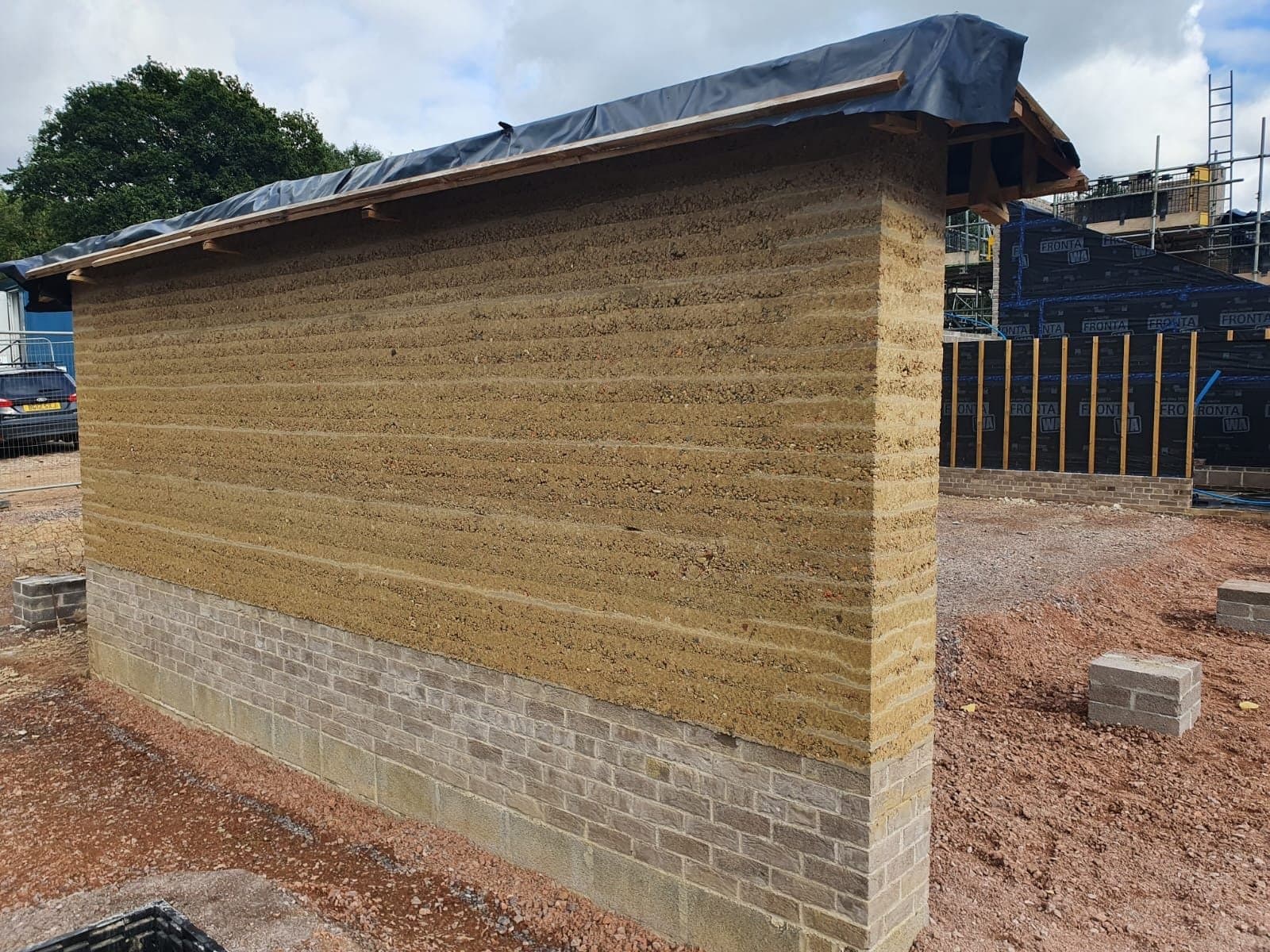 Struck rammed earth wall on a brick plinth showing its layers, under black protective sheeting