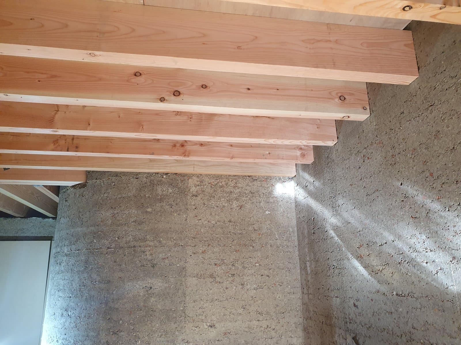 Rammed earth wall below timber floor joists inside the finished house, with light across the face