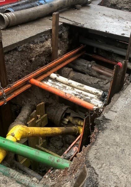 Trench sheets and a waler frame with orange hydraulic struts spanning an excavation crossed by existing services